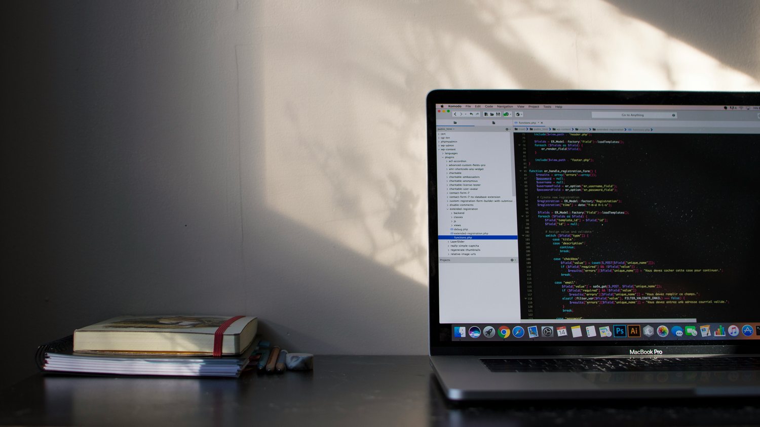 A laptop showing a code editor beside a stack of notebooks on a dark desk, leaf shadows falling on the wall behind