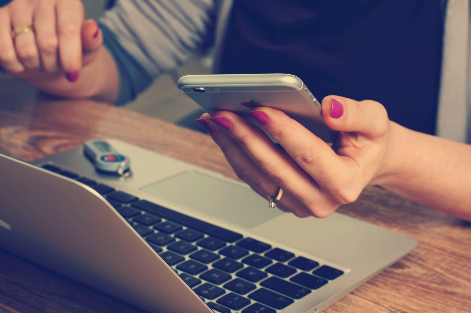 A pair of hands holding a phone above an open laptop on a wooden table, a second person seated opposite