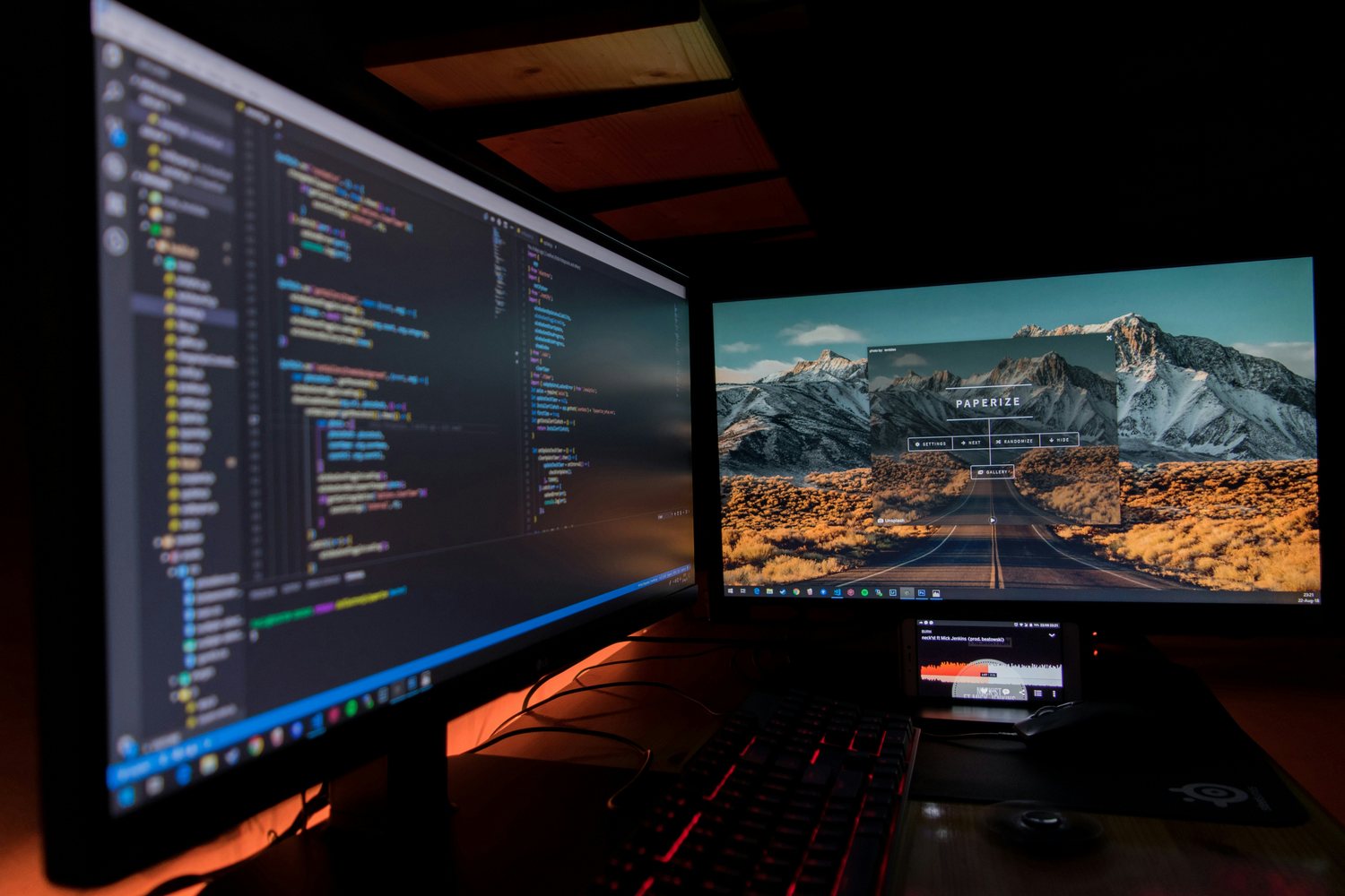 Two monitors in a darkened room, one filled with code and one showing a desert landscape, above a red-lit keyboard