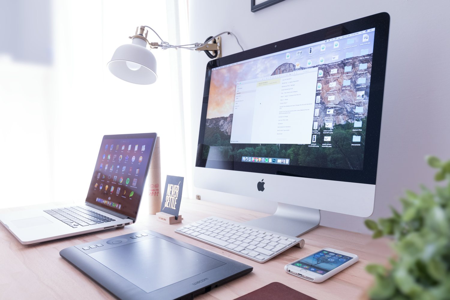 A sunlit desk holding a large desktop monitor, an open laptop, a graphics tablet and an angled reading lamp