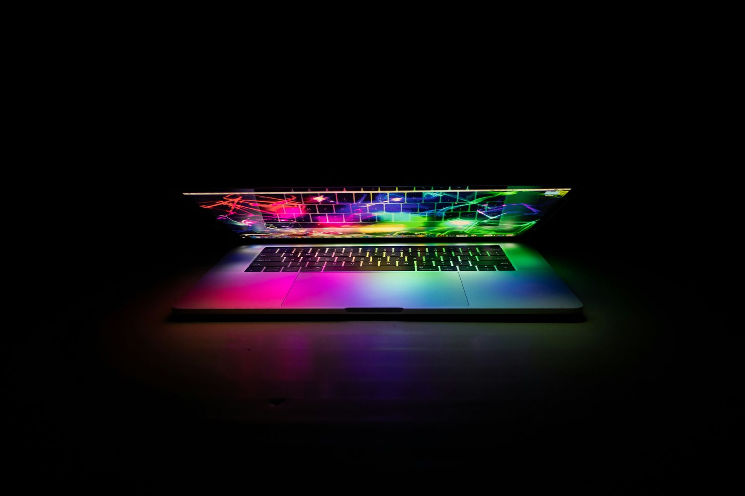 An open laptop in a darkened room lit by a spectrum of colour from its screen