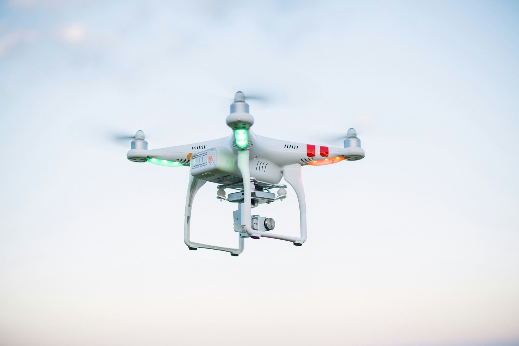 A white quadcopter drone with a gimbal-mounted camera hovering against a pale sky
