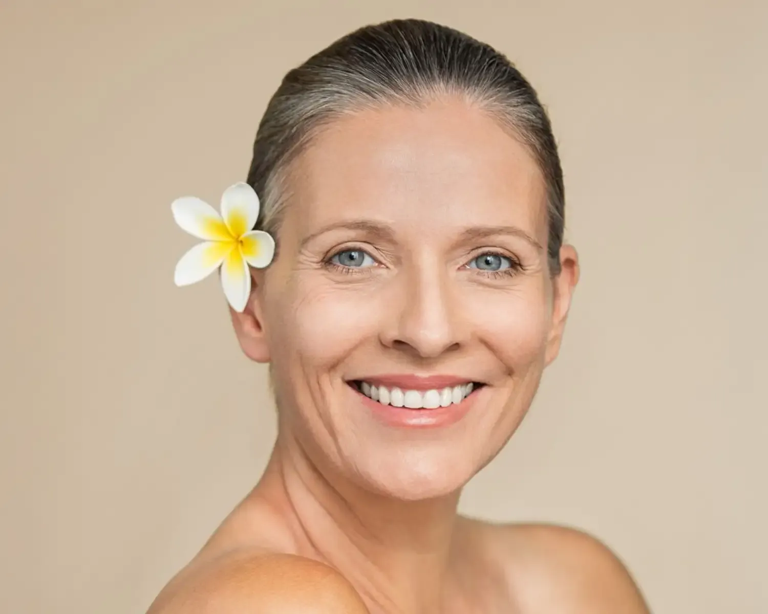 Mujer sonriente con flor blanca en el cabello, sobre fondo beige.